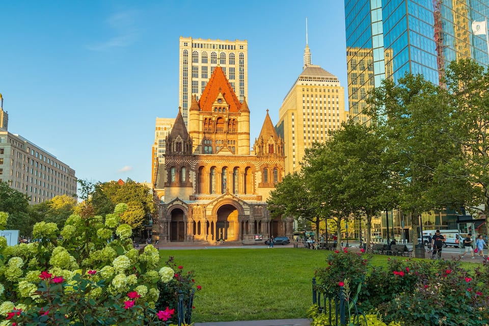 A historic church with a pointed roof and a large tower, surrounded by modern buildings and a well-maintained park with flowers and trees.