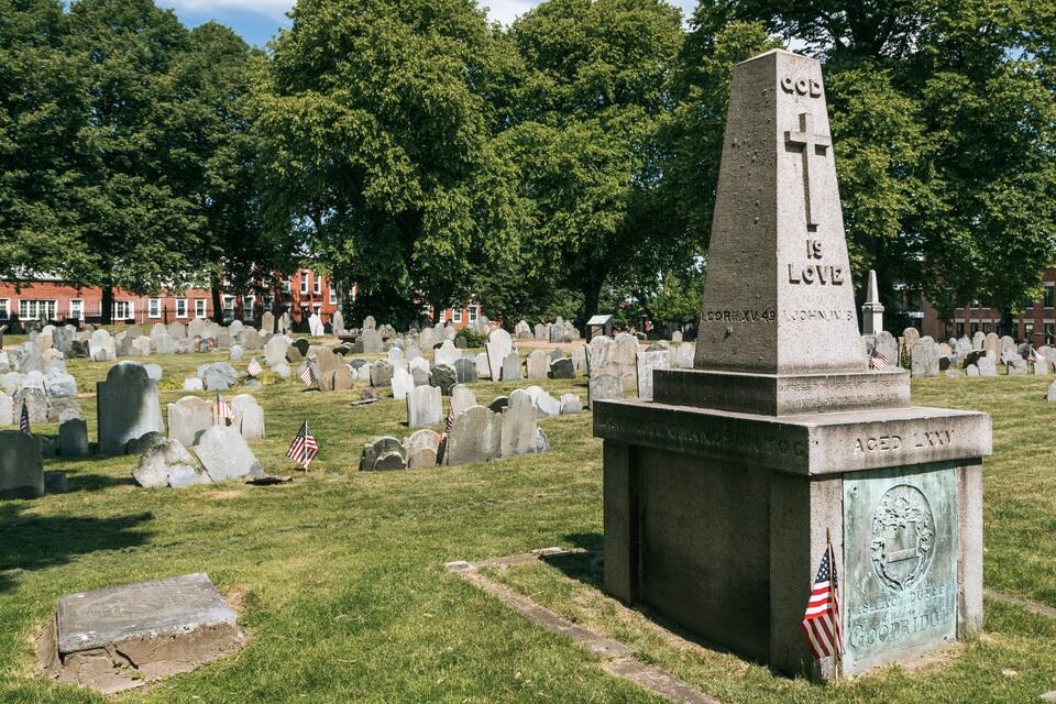Un cementerio con numerosas lápidas, un monumento destacado con inscripciones y banderas estadounidenses colocadas sobre algunas de ellas.