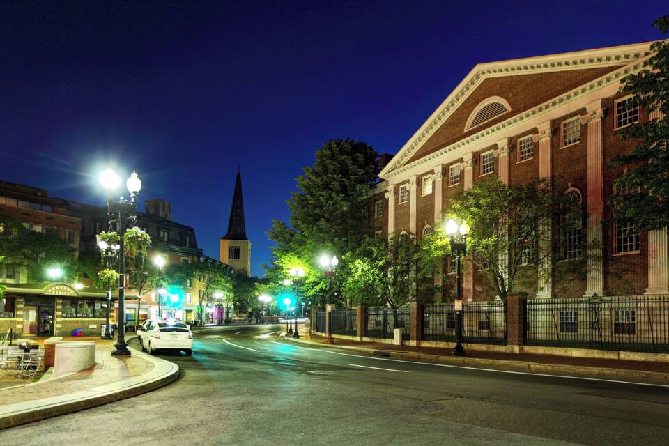 A nighttime street scene with a historic building, street lamps, and a church spire.