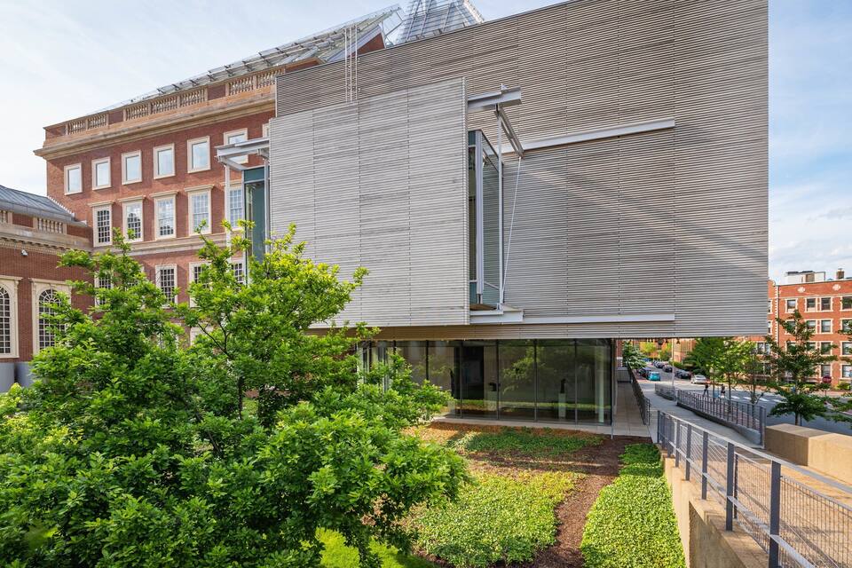 A modern building with a glass facade, surrounded by lush greenery and adjacent historic buildings.