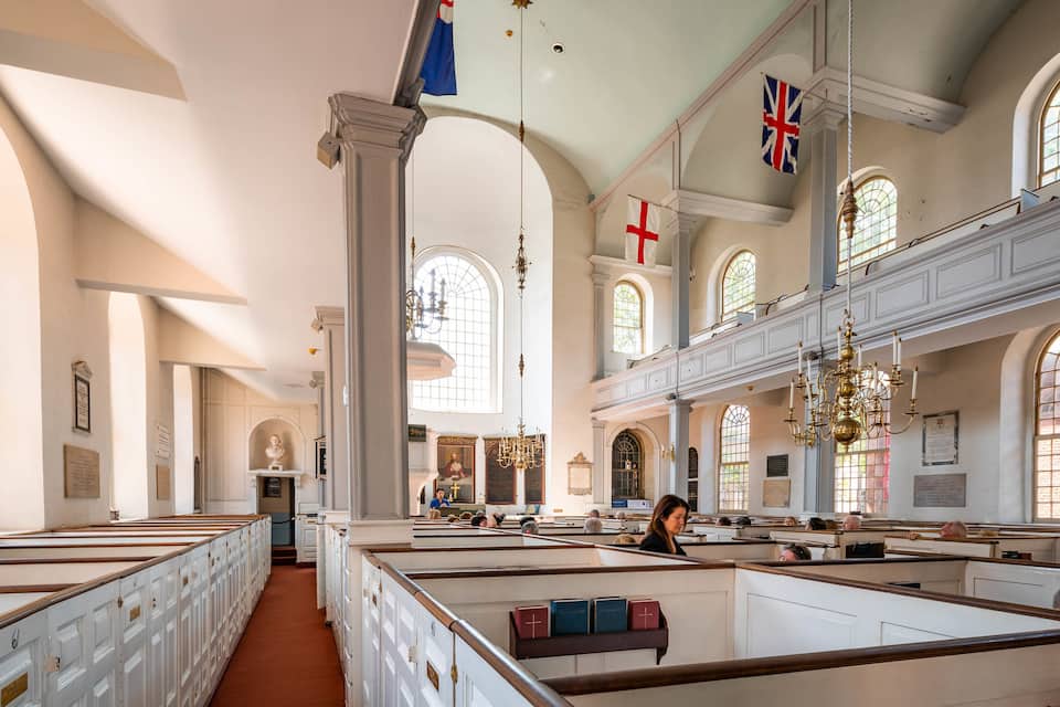 Interior de igreja com bancos, vitrais e bandeiras.