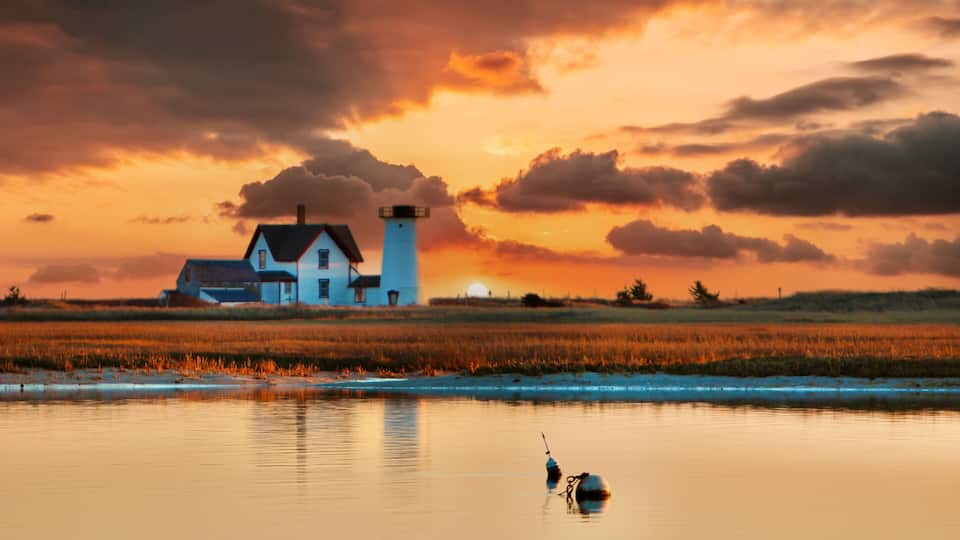 A lighthouse, a house, and a calm water body during sunset.