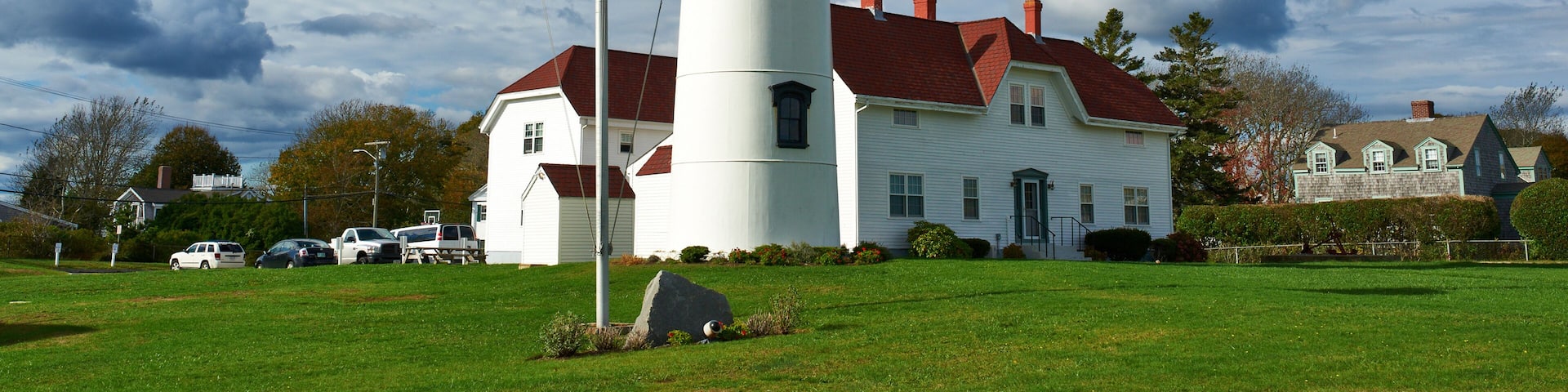 A lighthouse with an American flag, a white building with a red roof, and a cloudy sky.