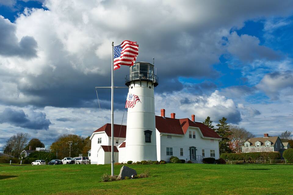 Um farol com a bandeira americana, um edifício branco com telhado vermelho, e um céu nublado.