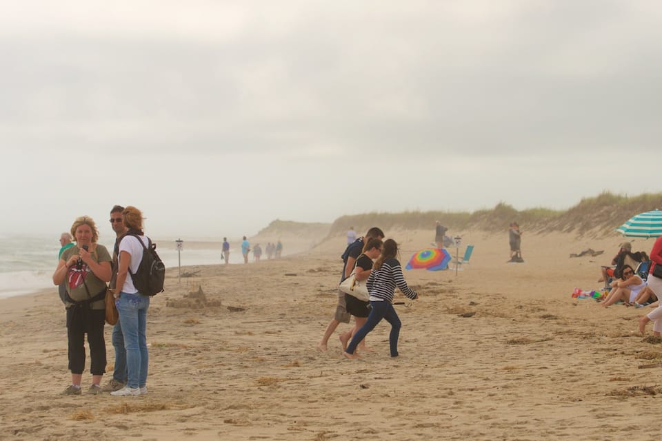 Una playa con gente caminando y de pie, un castillo de arena y un paraguas de playa a rayas.