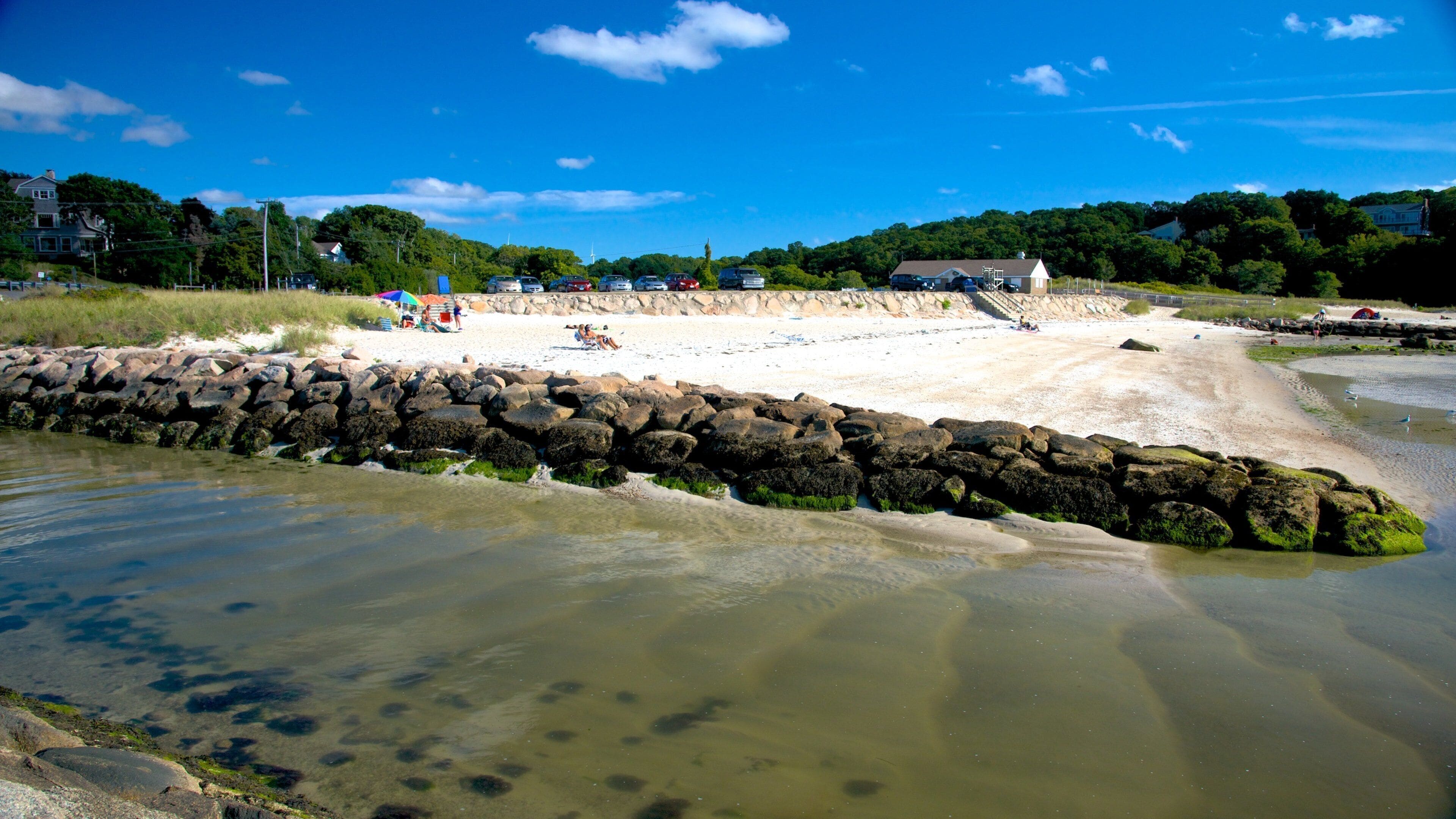 A sandy beach with a rocky breakwater, clear water, and houses in the background.