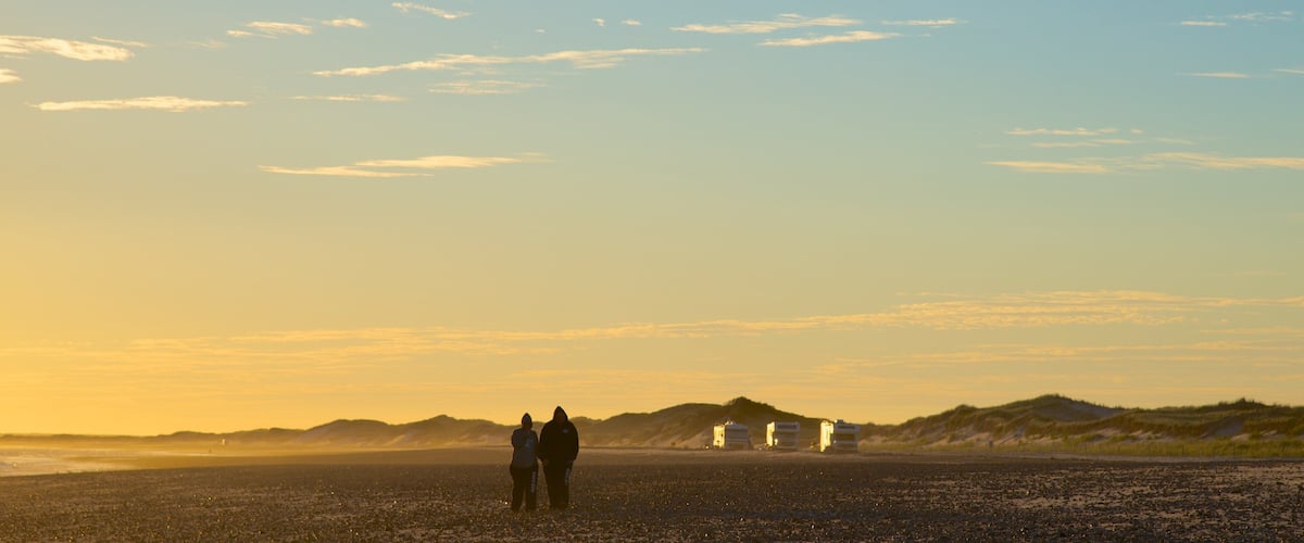 A beach with people standing, a dune, and a building in the distance.