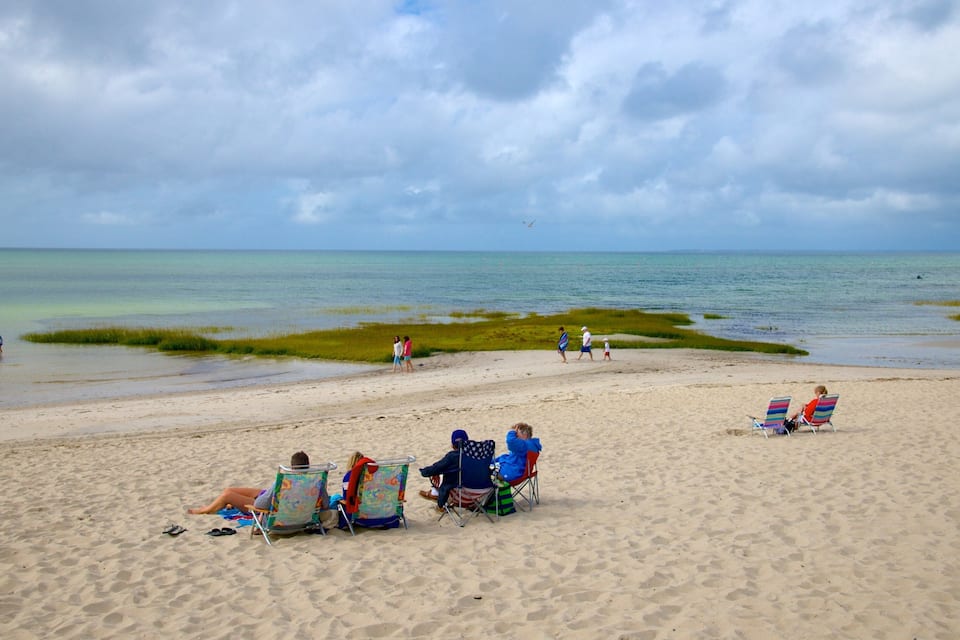 Una playa arenosa con gente descansando en sillas de playa coloridas, un mar tranquilo y un cielo nublado.