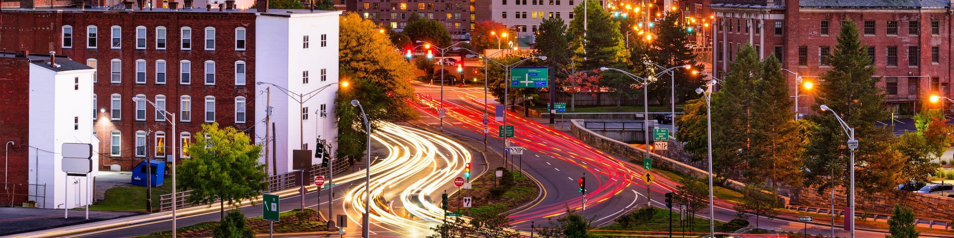 A cityscape at dusk with illuminated buildings, a prominent church spire, and a busy intersection with light trails from moving vehicles.