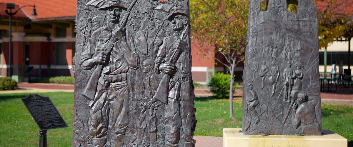 Two large bronze sculptures of soldiers in combat poses, set against a backdrop of a red brick building and autumn-colored trees.