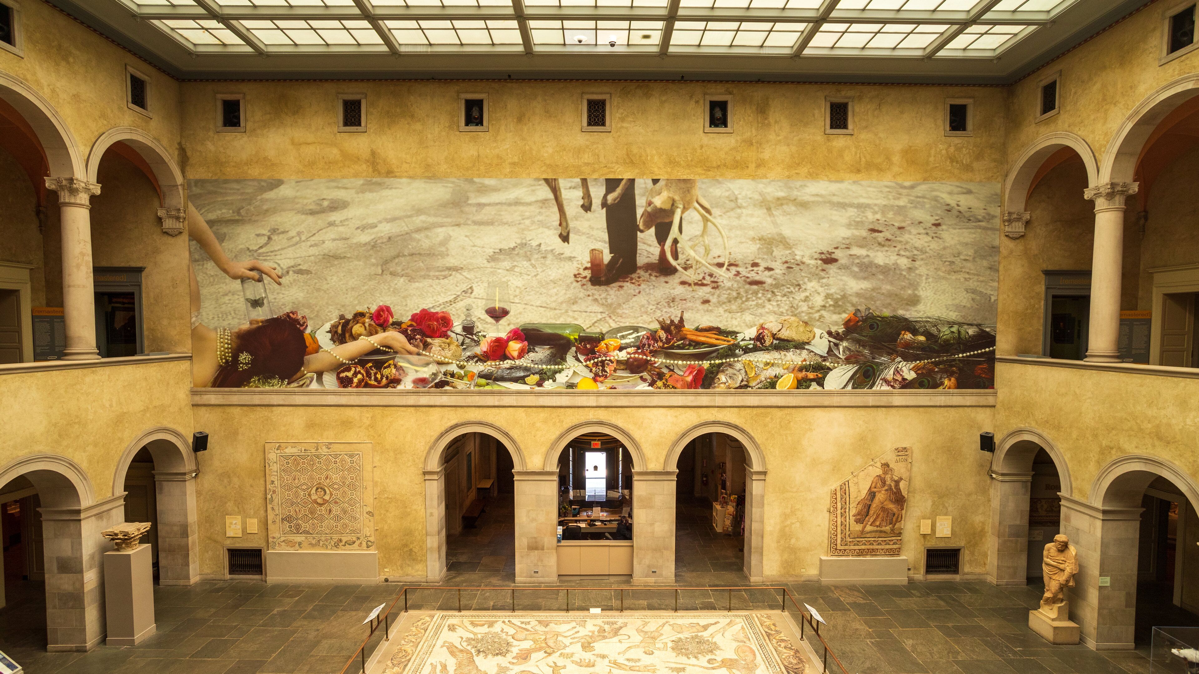 A grand hall with a large mural depicting a historical scene, arched columns, and a central statue.