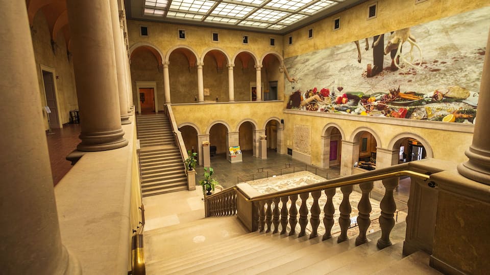 A grand interior with a central staircase, arched columns, and a detailed mural on the wall.