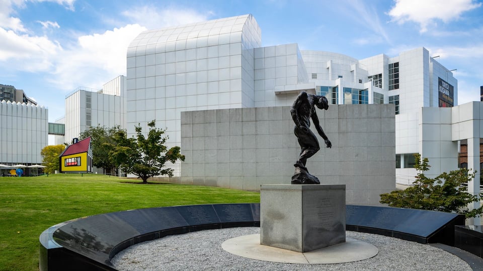 A modern building with a glass facade, a sculpture of a man, and a small yellow house in the foreground.