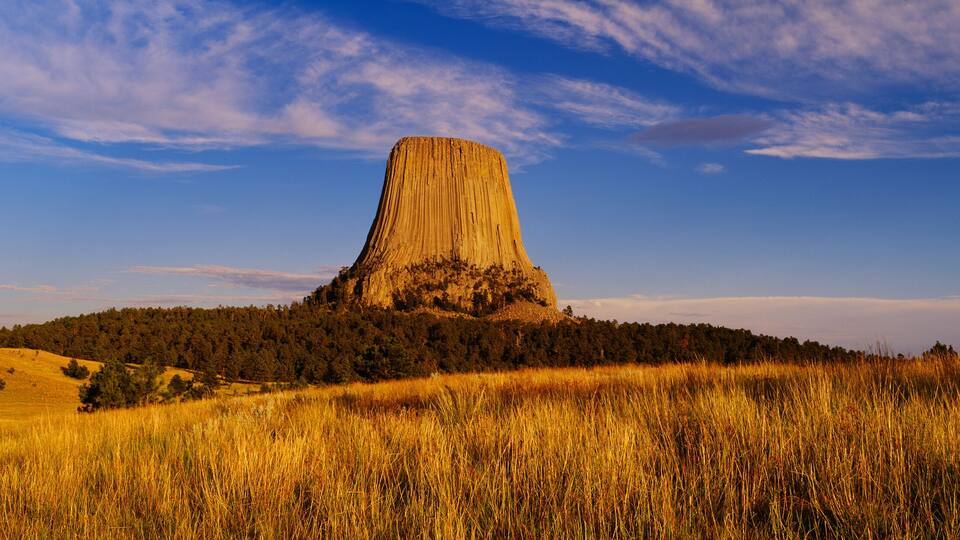 A tall, conical rock formation surrounded by a forested hill and golden grass.