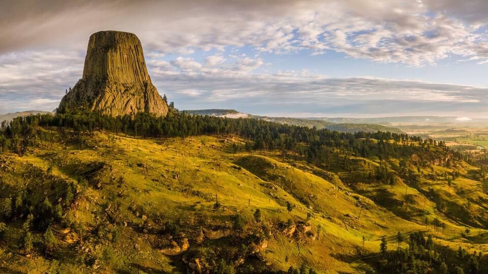 A large, steep rock formation surrounded by forested hills and a winding road.