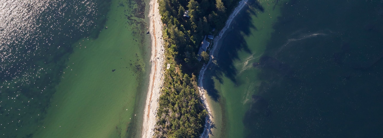 Vista aérea de una zona costera con una línea de costa curva, agua verdosa y una estrecha franja de tierra con edificios dispersos.