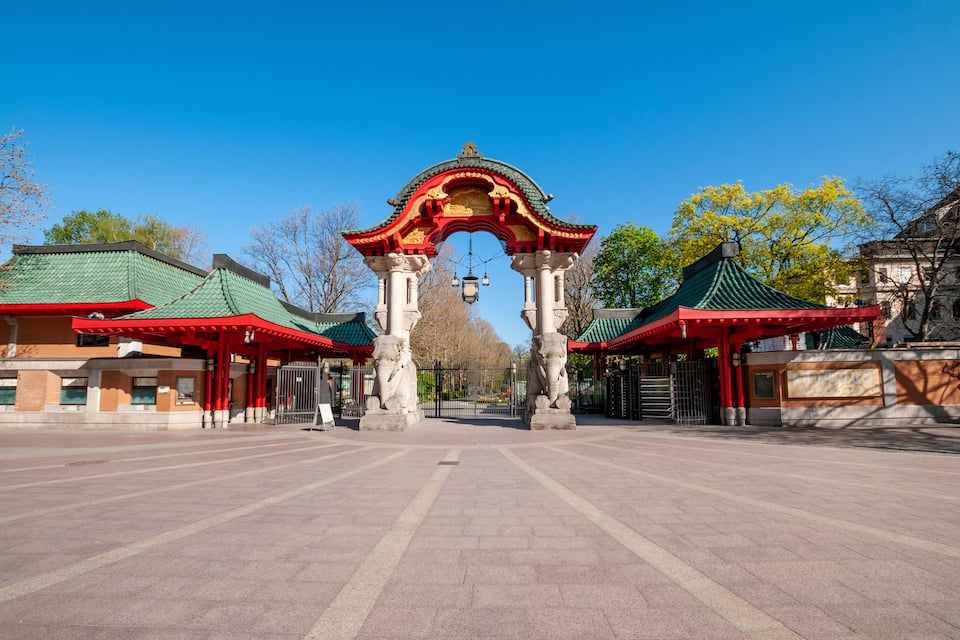 Una puerta china tradicional con techos rojos y verdes, flanqueada por estatuas de leones de piedra, que da a un patio con suelo de baldosas.