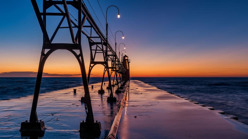 A coastal bridge at sunset with a calm sea and street lamps.
