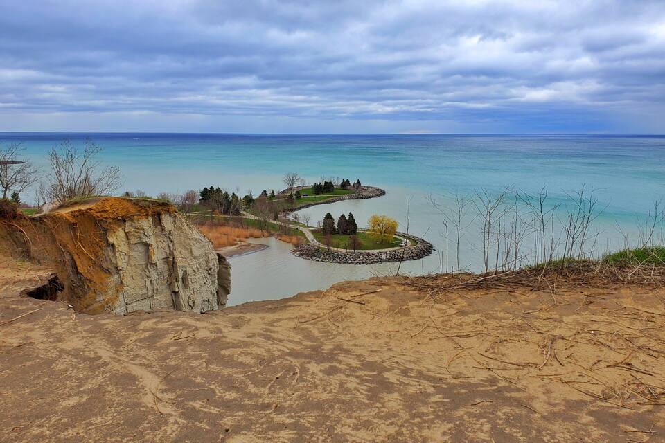 Eine Küstenlandschaft mit einem felsigen Klippenhang, einem sandigen Vordergrund und einer ruhigen Bucht mit klarem, blauem Wasser.