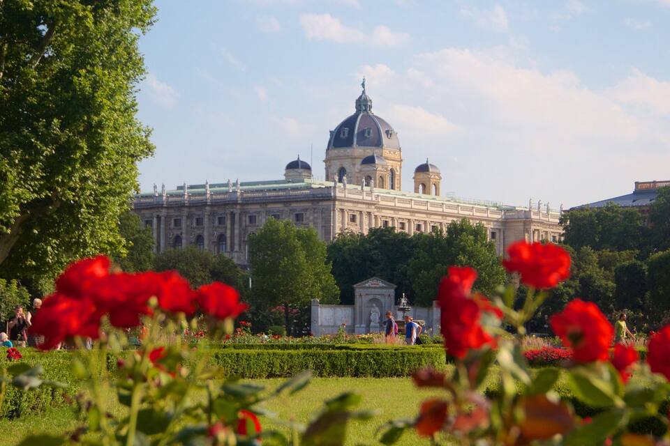 A large classical building with domes and statues, surrounded by lush greenery and red flowers.