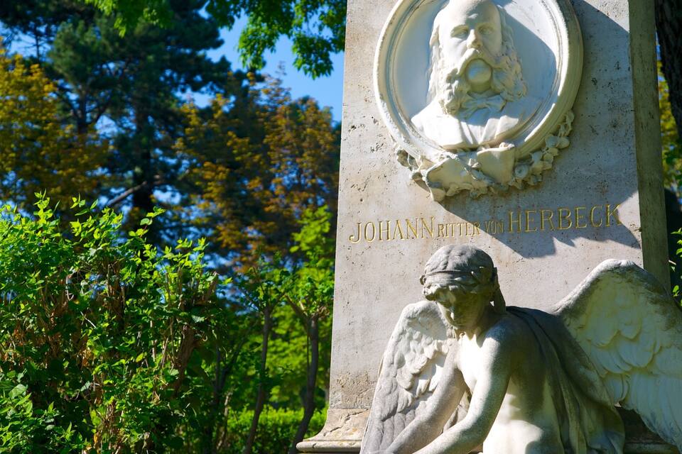 A gravestone with a bust and an angel statue, surrounded by greenery.