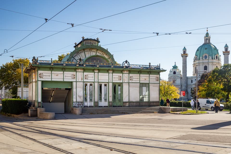 A tram station with green and white architecture, a tram track, and a large church with a green dome in the background.