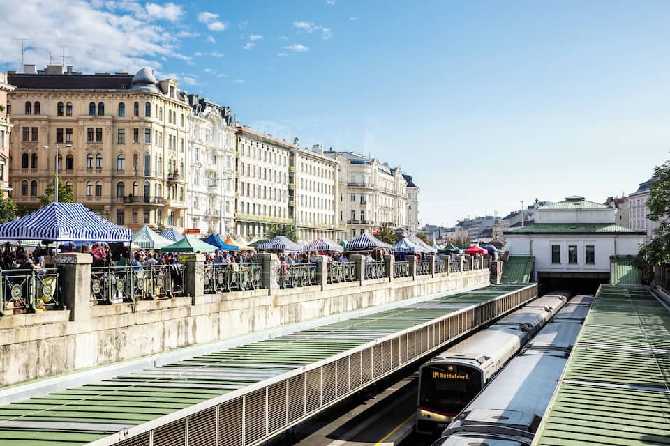 A train station with a green roof, a train on the tracks, and a crowd of people under striped umbrellas.
