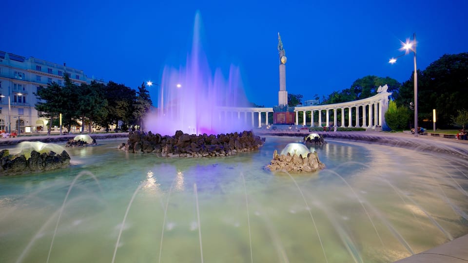 A fountain with water jets and illuminated by purple lights, surrounded by a plaza with a tall monument and classical architecture.
