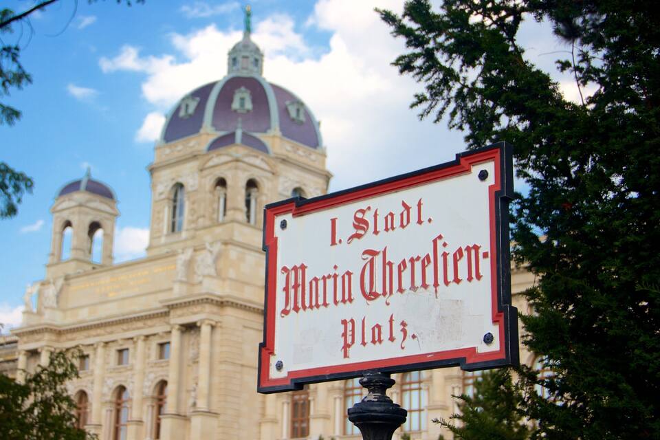 A sign in front of a large building with a dome and two smaller domes, reading "I. Stadt Maria Therese Platz."