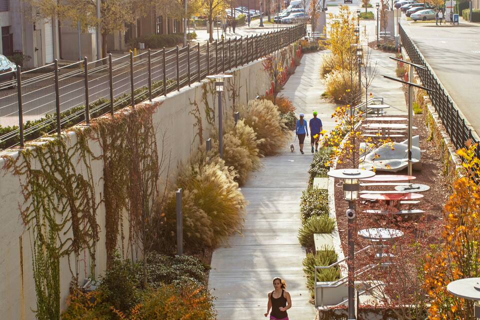 Un paseo peatonal bordeado de árboles y plantas, una cerca de metal y un cartel que dice 'Macy City Park'.