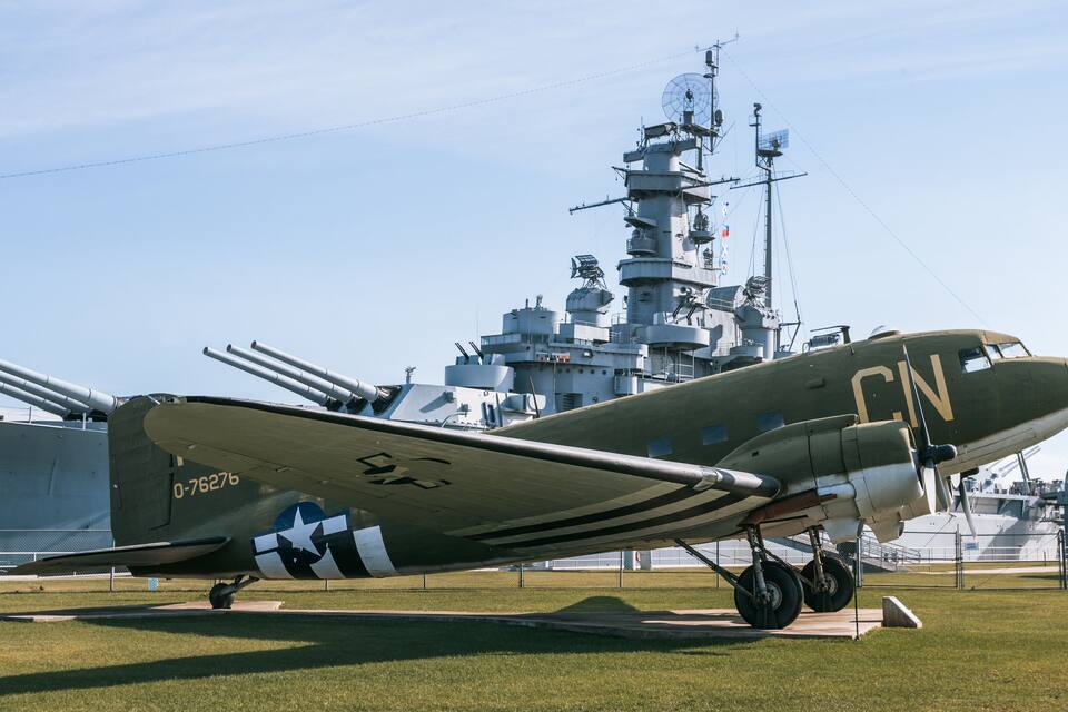 Un avión militar con el número 0-76276 en la cola, estacionado en un campo cubierto de hierba con un buque naval al fondo.