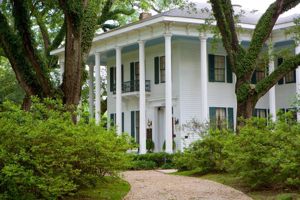 A white colonial-style house with green shutters, surrounded by lush greenery and trees.