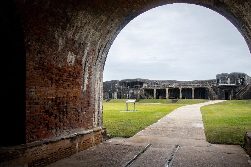 Une structure en briques en forme d’arche mène à un bâtiment historique doté d’une cour verdoyante et d’une enseigne.