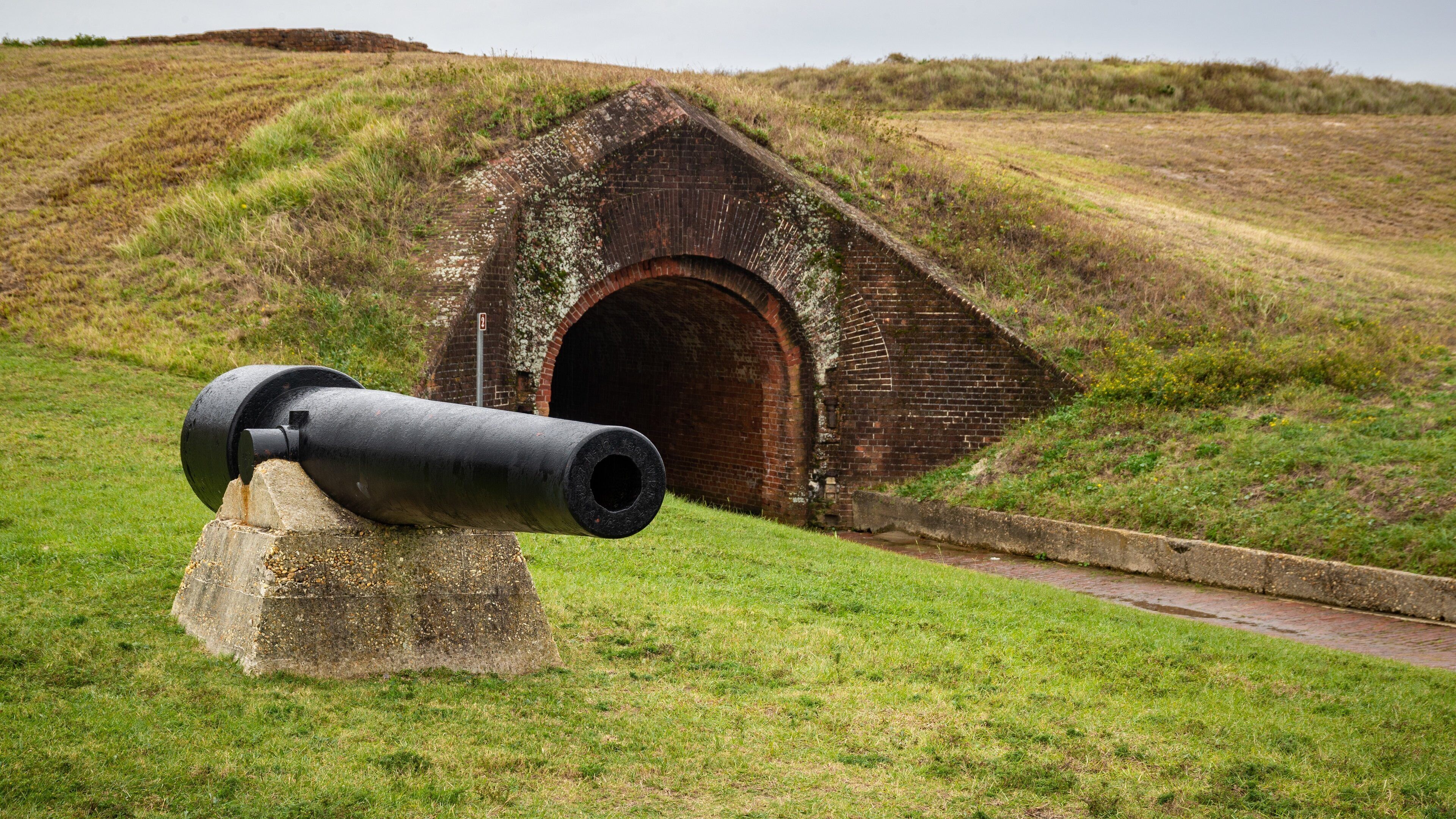 A cannon on a grassy hill with a brick structure in the background.
