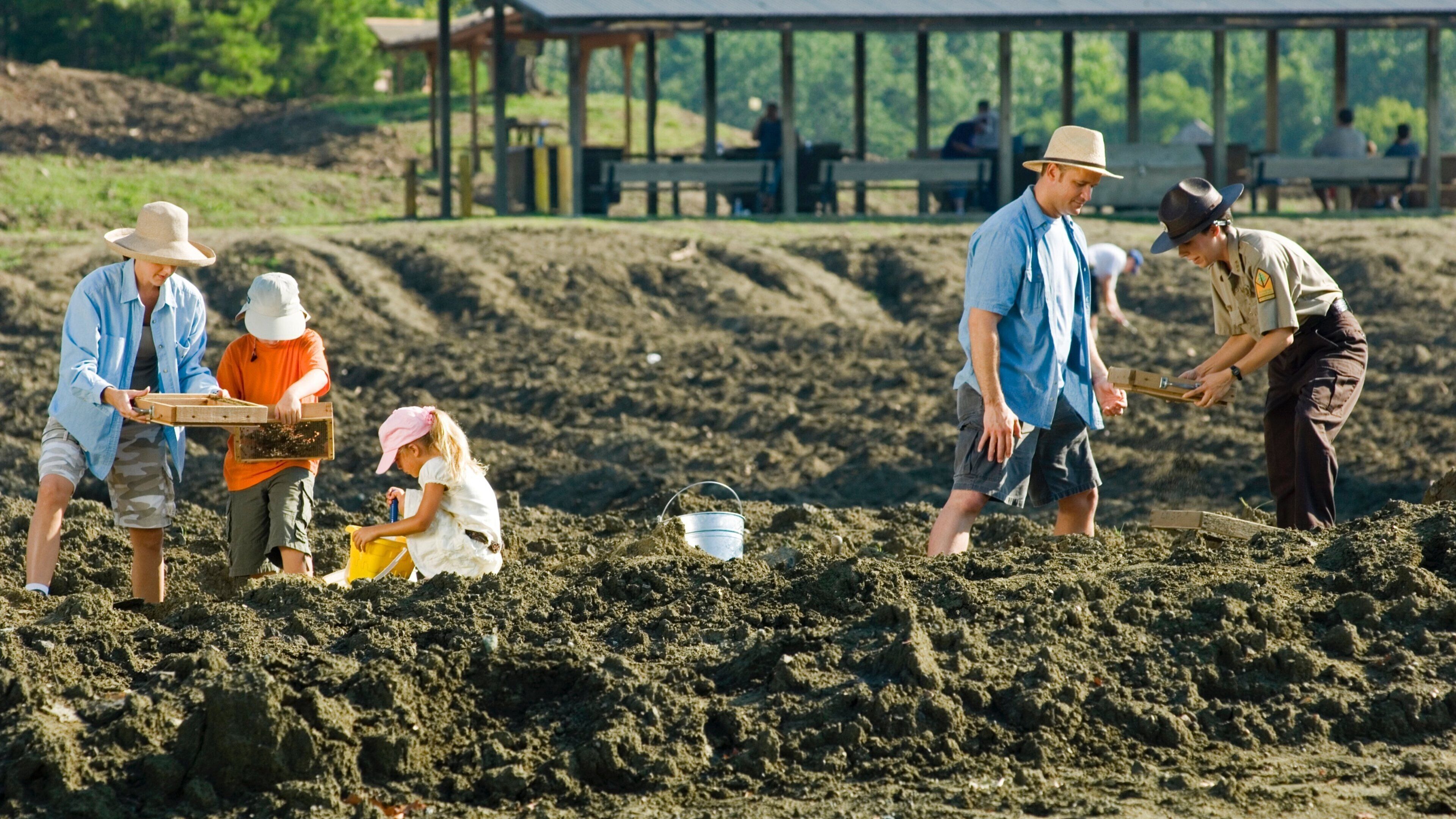 People working in a sandy field with a wooden structure in the background.