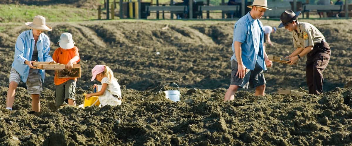 People working in a sandy field with a wooden structure in the background.