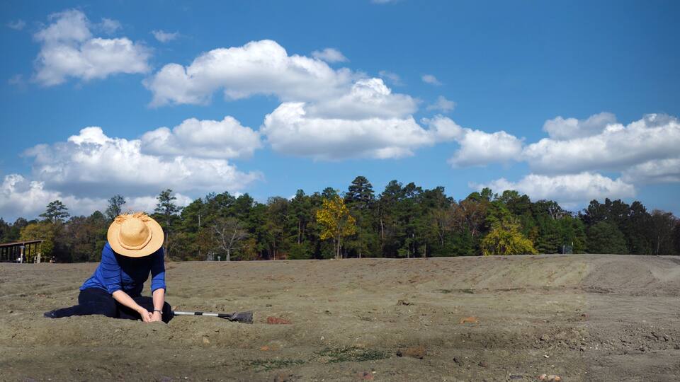 A person in blue attire and a straw hat working on a dirt field with a rake, under a clear blue sky with scattered clouds, and a forested background.