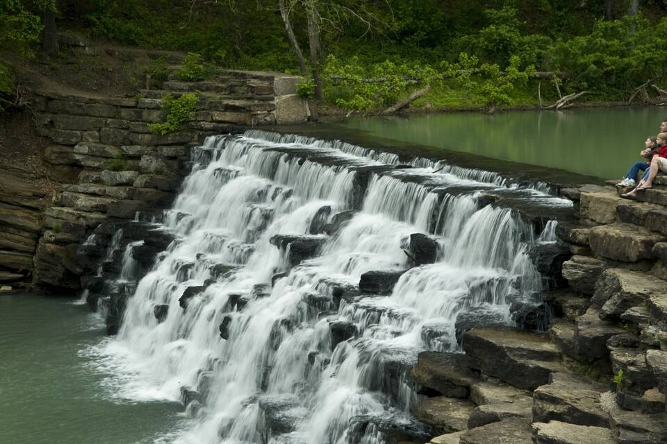 A multi-tiered waterfall cascading over rocks into a calm body of water, surrounded by lush greenery.
