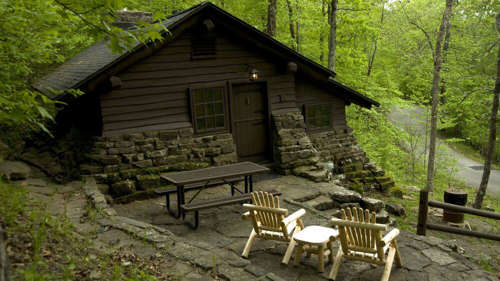 A rustic cabin with a stone foundation, wooden walls, and a chimney. There is a picnic table and two Adirondack chairs in front of the cabin.