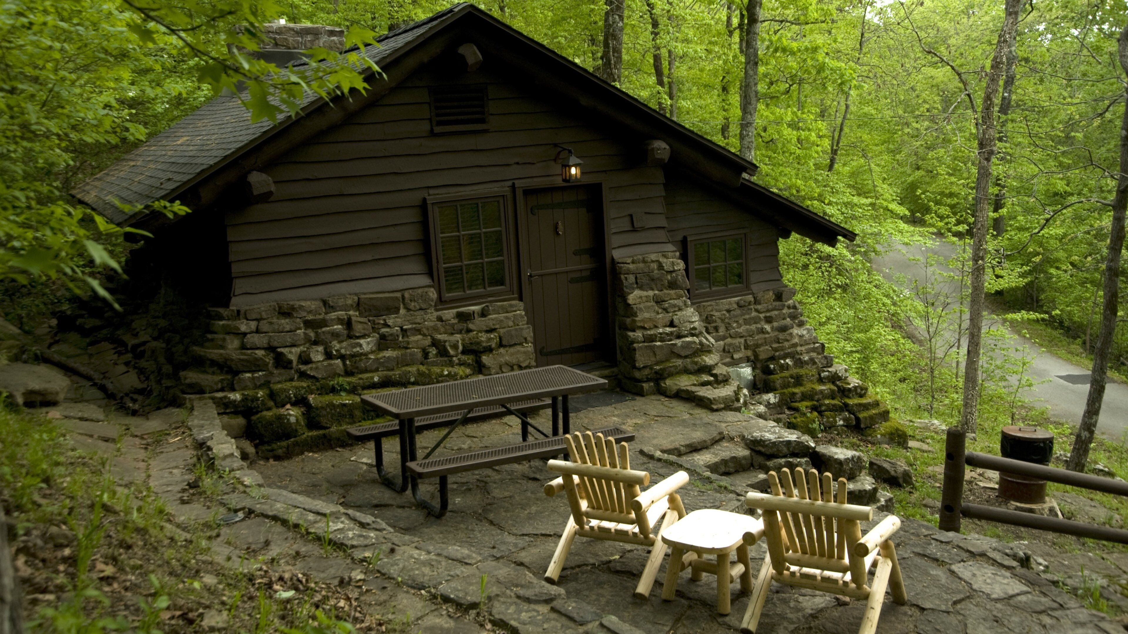 A rustic cabin with a stone foundation, wooden walls, and a chimney. There is a picnic table and two Adirondack chairs in front of the cabin.