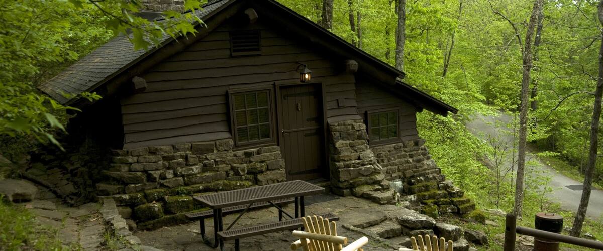 A rustic cabin with a stone foundation, wooden walls, and a chimney. There is a picnic table and two Adirondack chairs in front of the cabin.