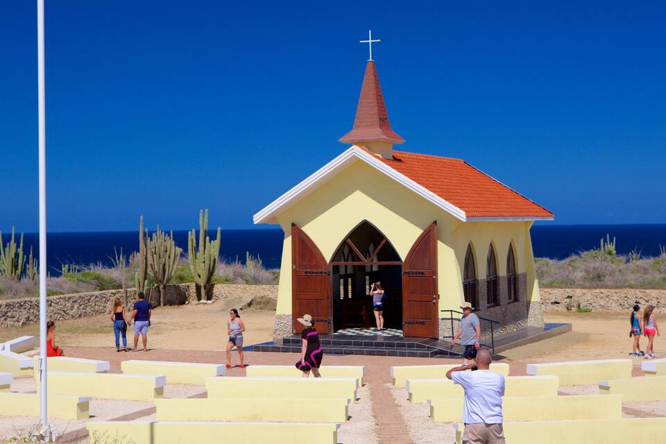 Una iglesia pequeña con techo rojo y cruz en la parte superior, cerca de una playa con cactus y el océano al fondo.
