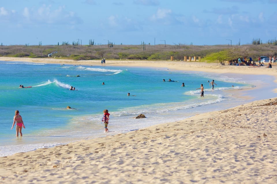 Una playa con agua turquesa cristalina, gente nadando y practicando surf, y una orilla de arena con vegetación escasa.