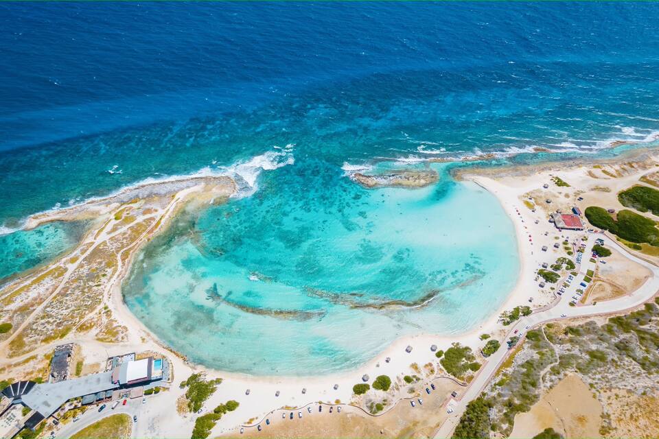 An aerial view of a coastal area with a sandy beach, clear turquoise waters, and a small building with a red roof.