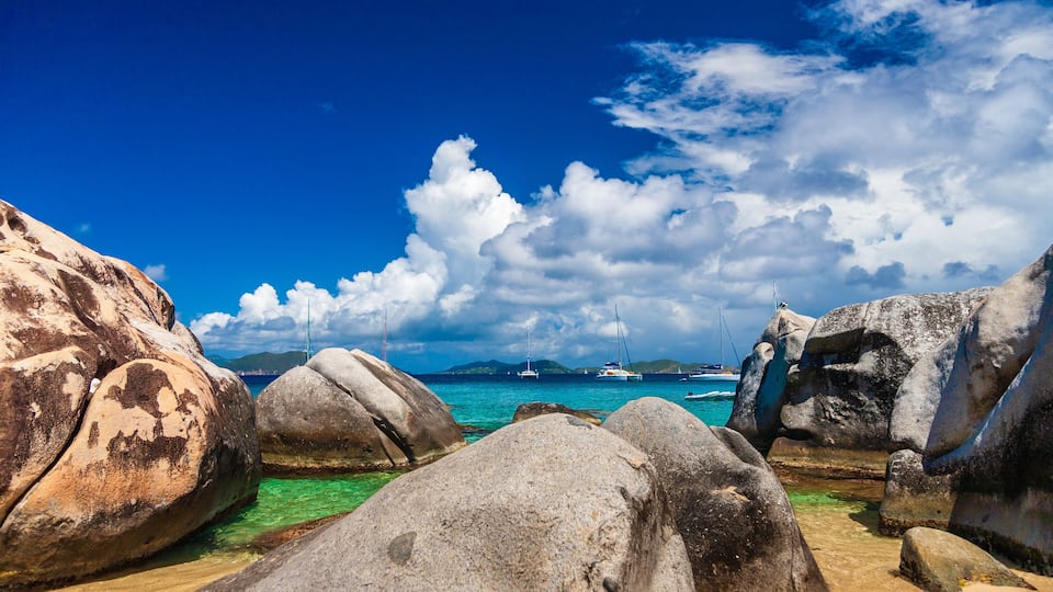 A rocky beach with clear turquoise water, large boulders, and sailboats in the distance.