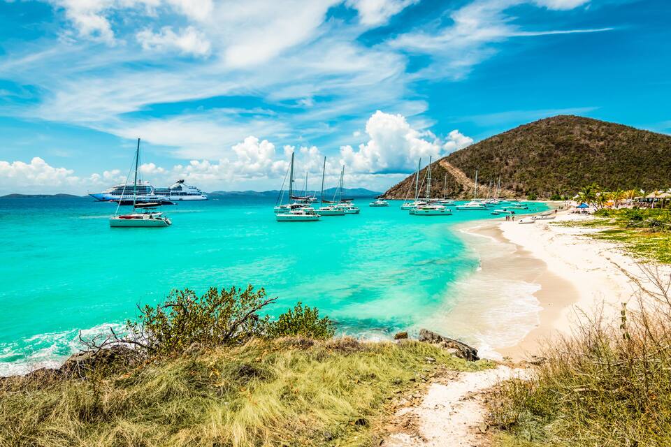 Una playa turquesa con veleros, un crucero y un telón de fondo montañoso.