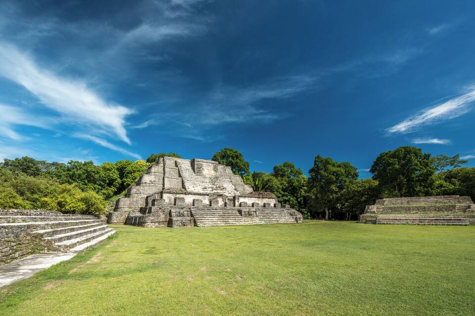 A large ancient Mayan pyramid with a stepped structure, surrounded by lush green grass and trees under a clear blue sky.