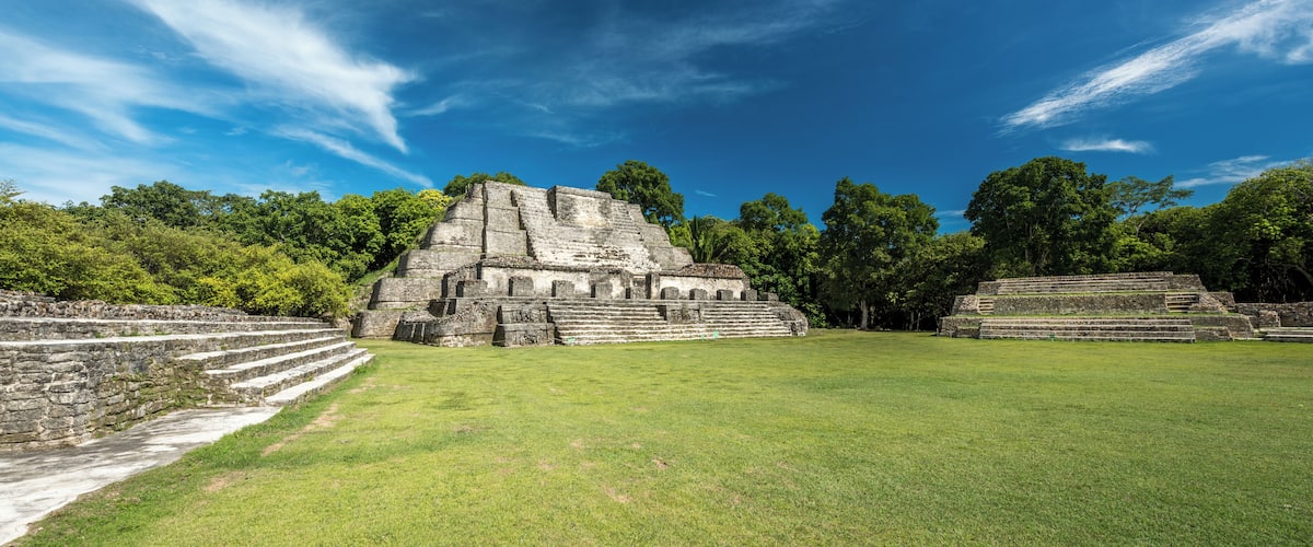 A large ancient Mayan pyramid with a stepped structure, surrounded by lush green grass and trees under a clear blue sky.