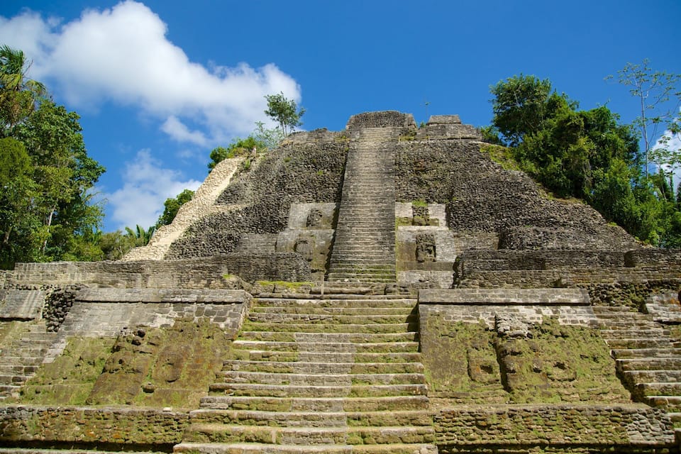 Une pyramide de pierre, avec un escalier menant à son sommet, entourée d’une végétation luxuriante et sous un ciel bleu clair.