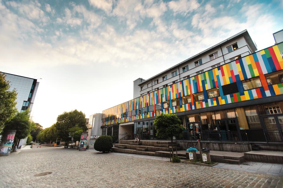 Um edifício colorido com design moderno, rua de paralelepípedos e árvores ao fundo.